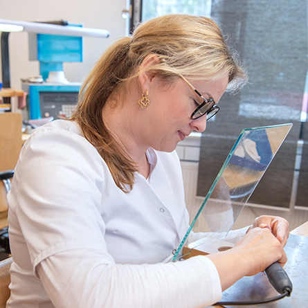 Zahntechnikerin schleift an einem Objekt hinter einer Glasscheibe.