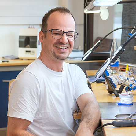 Lächelnder Zahntechniker mit Brille sitzt am Arbeitsplatz im Dentallabor.