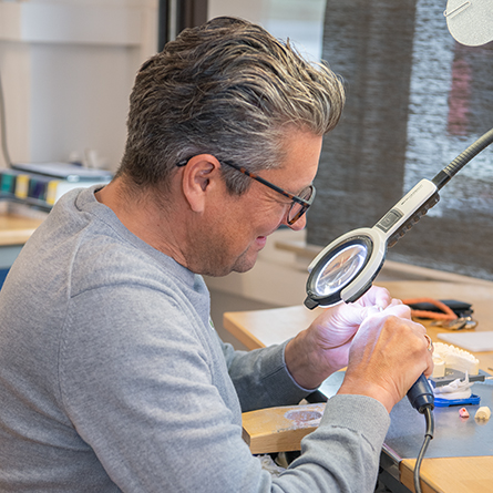 Zahntechniker bei präziser Handarbeit unter Lupenleuchte, Zahnmodell in der Hand.