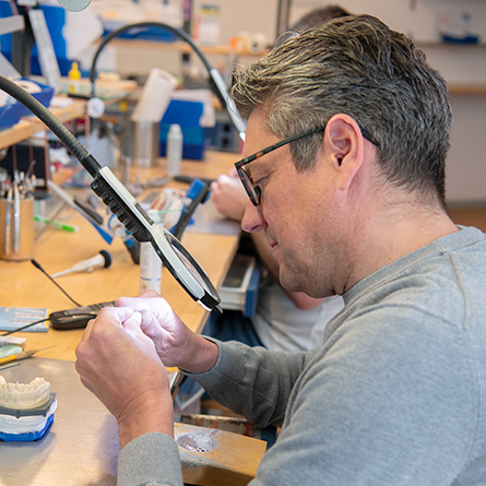 Dentaltechniker mit Brille arbeitet konzentriert mit einer Lupe an einem Zahntechnikmodell.