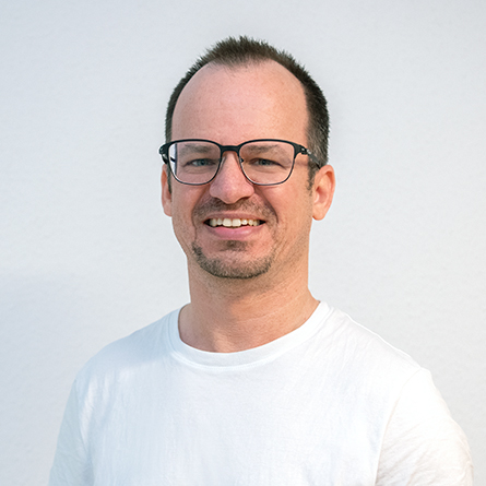 Porträt eines Mannes mit Brille und weißem T-Shirt, lächelnd vor hellem Hintergrund.