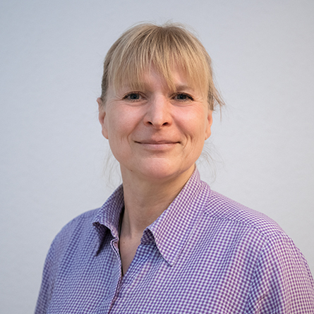 Porträt einer Frau mit blonder Ponyfrisur und kariertem Hemd, freundlich lächelnd.