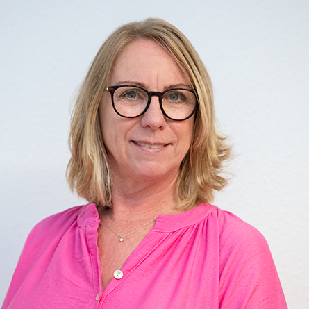 Mitarbeiterin mit blonder Bobfrisur, Brille und pinker Bluse vor hellem Hintergrund.